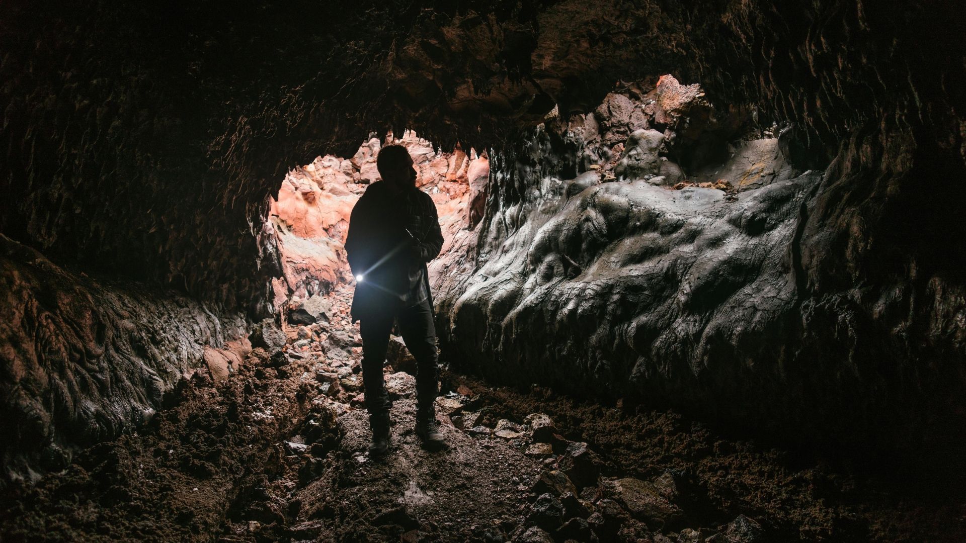 Person with flashlight in dark cave opening, illuminated by light.
