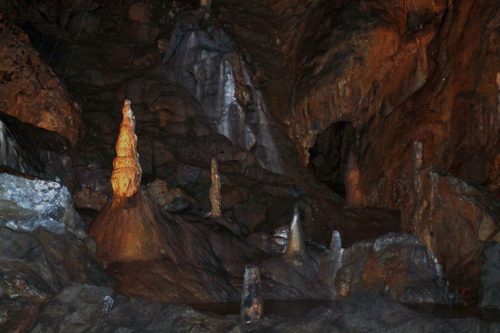 Inside a dark cave, brown stalactites and stalagmites are illuminated, creating an eerie, textured environment.