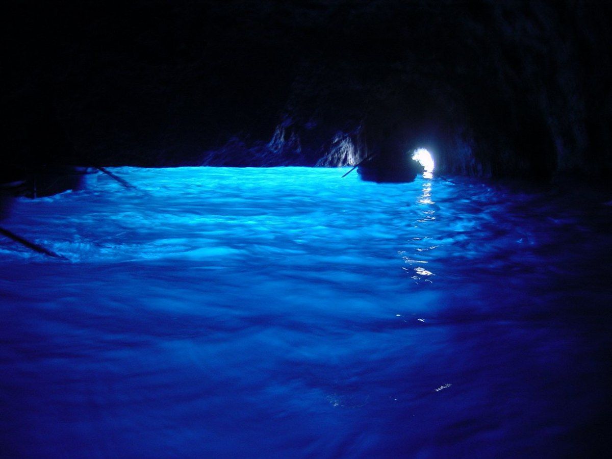 Inside a dark cave, bright blue water reflects light; a boat and natural opening are visible.
