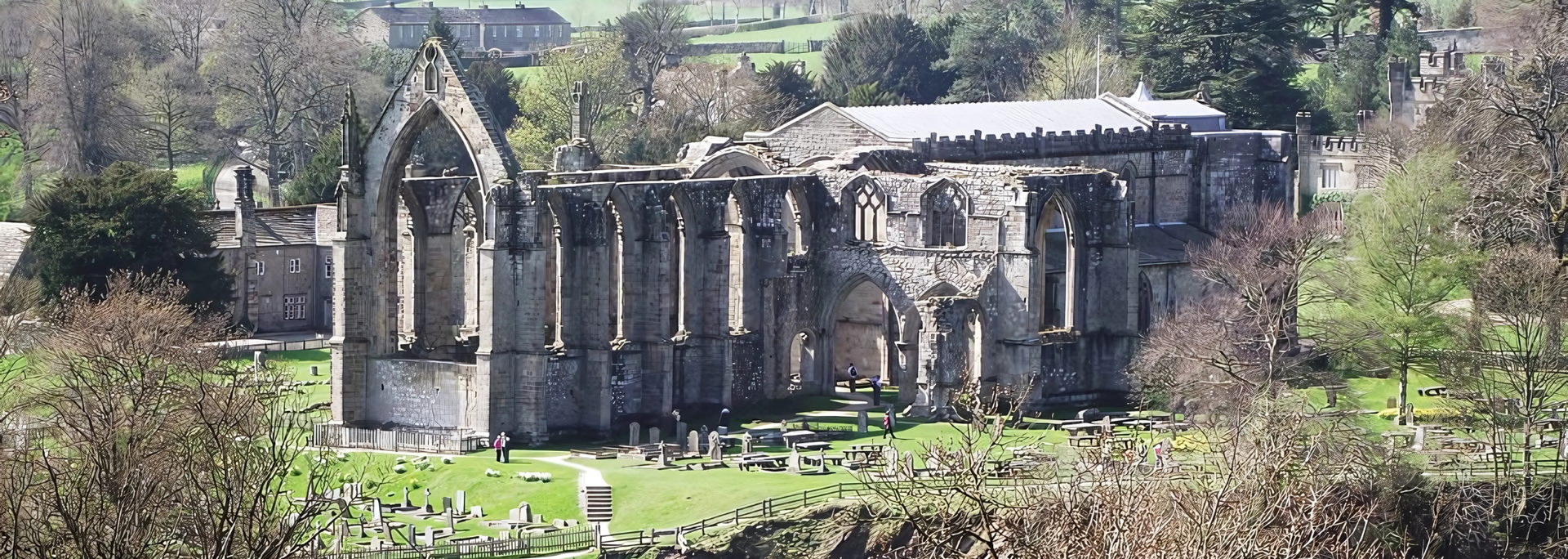 Ruined stone abbey in a green field with a cemetery and trees.