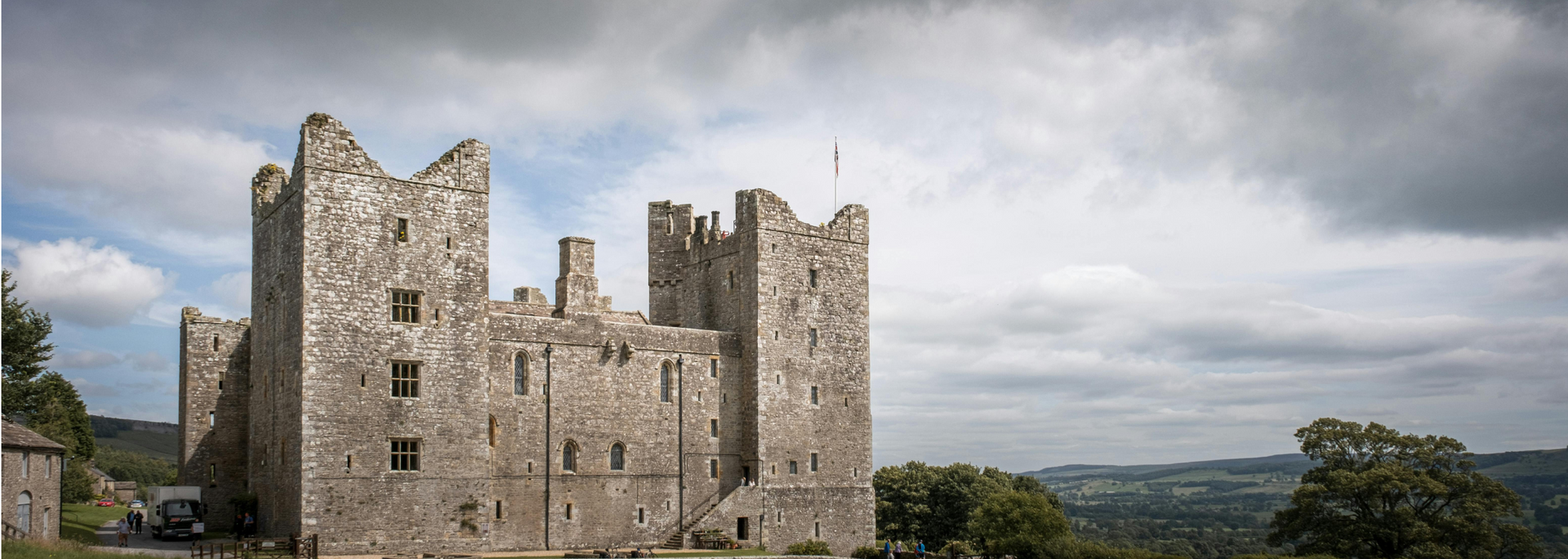 A weathered castle with stone towers sits on a hillside under a cloudy sky.