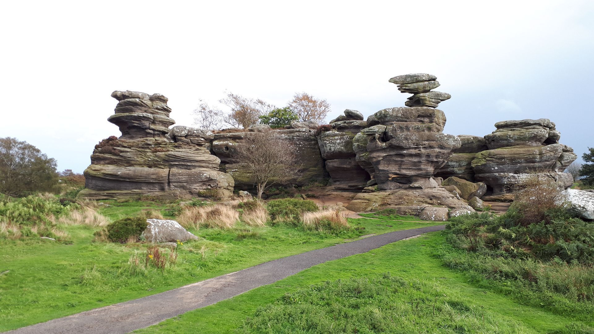 Rock formations in a grassy field with a paved path, under a cloudy sky.