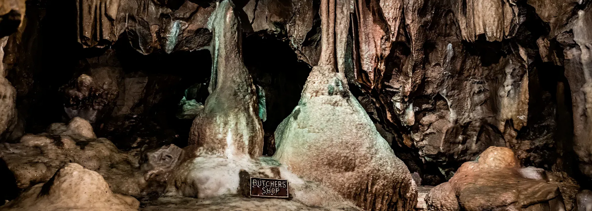 Inside a cave, stalactites hang from the ceiling. Stalagmites rise from the ground. Brown and tan rock formations.