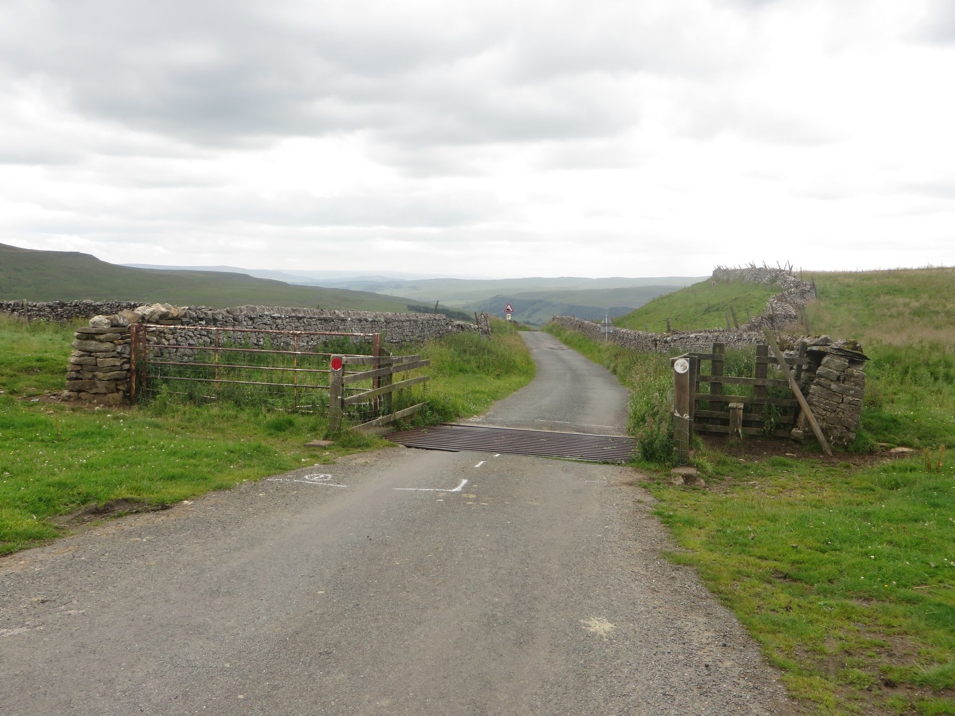 Gravel road through green field, passing a cattle grid. Stone walls flank the road, cloudy sky above.