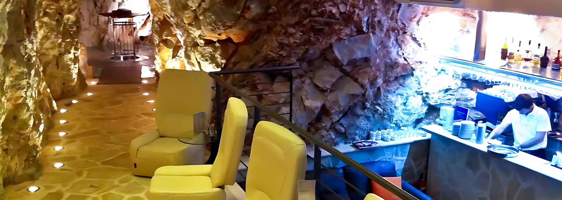 Cave-like interior with yellow chairs, a bar with a bartender, and a walkway lit by lights.