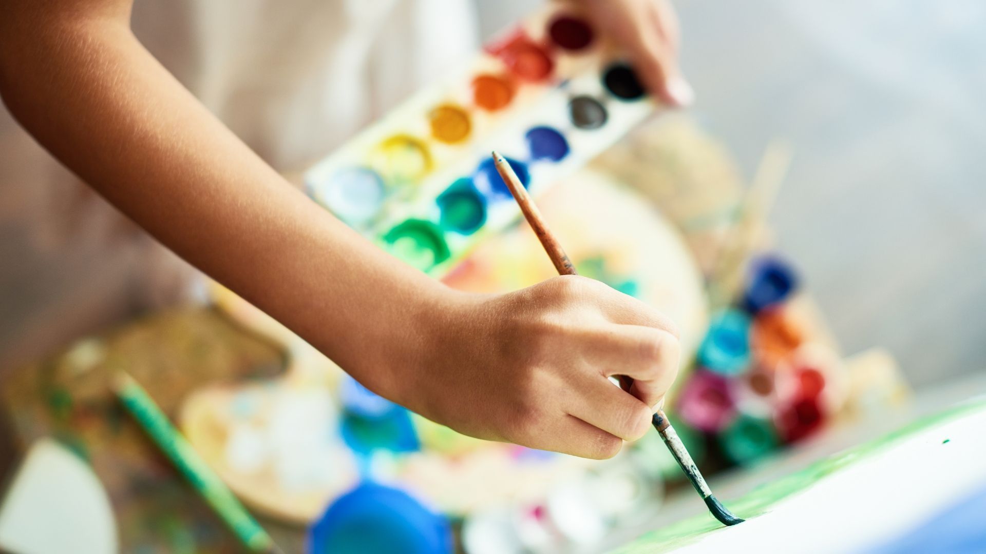 Child's hand holding a paintbrush, painting with watercolors from a palette on a table.