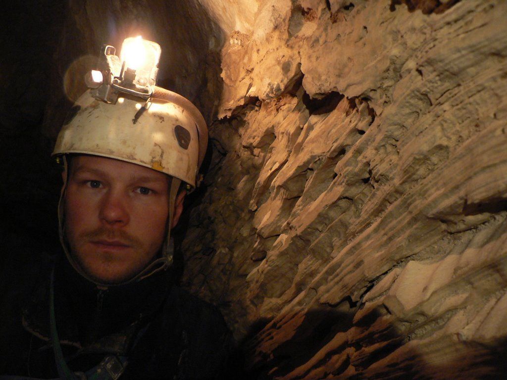 Man in helmet with headlamp in a cave, illuminated by light. Brown and beige rock walls.