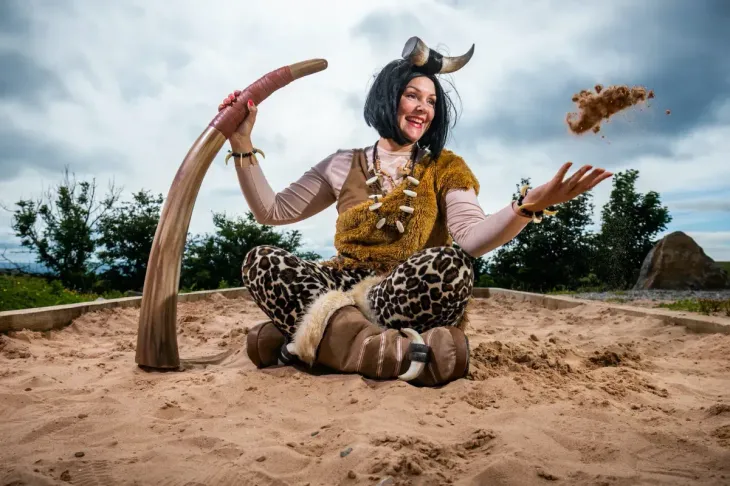 Woman in cavewoman costume sits in sand pit, juggling food and holding a tusk; cloudy sky.