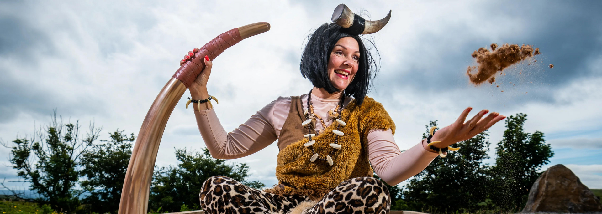 Woman in cavewoman attire throws a rock, holding a large curved object. Cloudy outdoor setting.