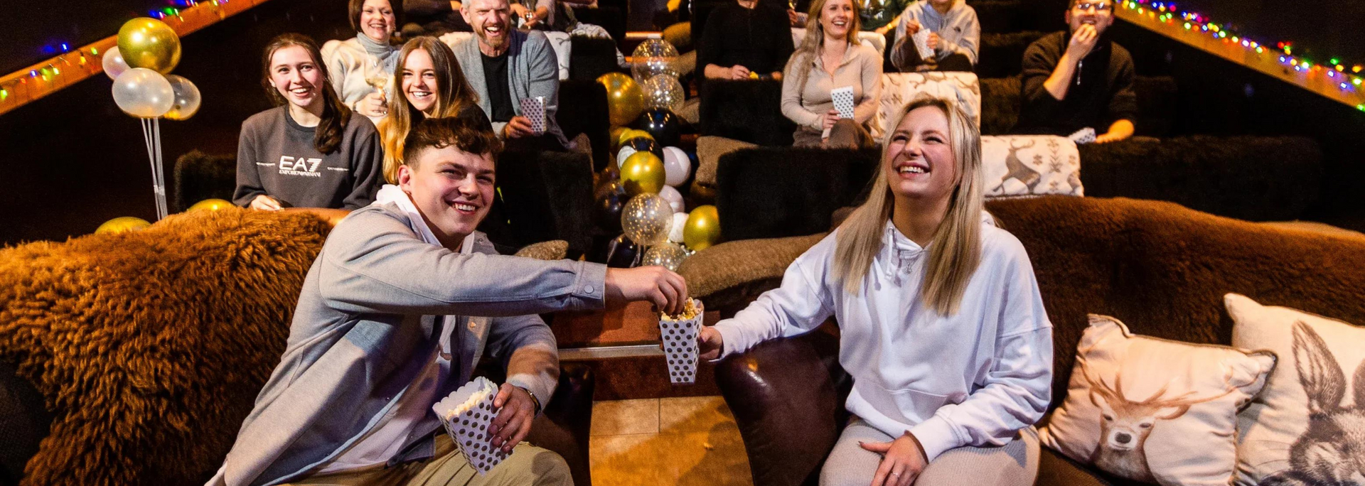 Two people holding cups, smiling at each other in front of an audience, party setting.