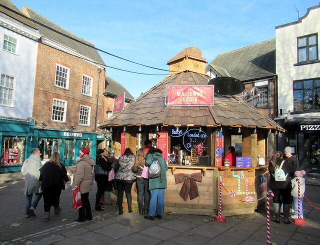 People queueing at a wooden kiosk selling food/drinks, set in a town square with buildings.