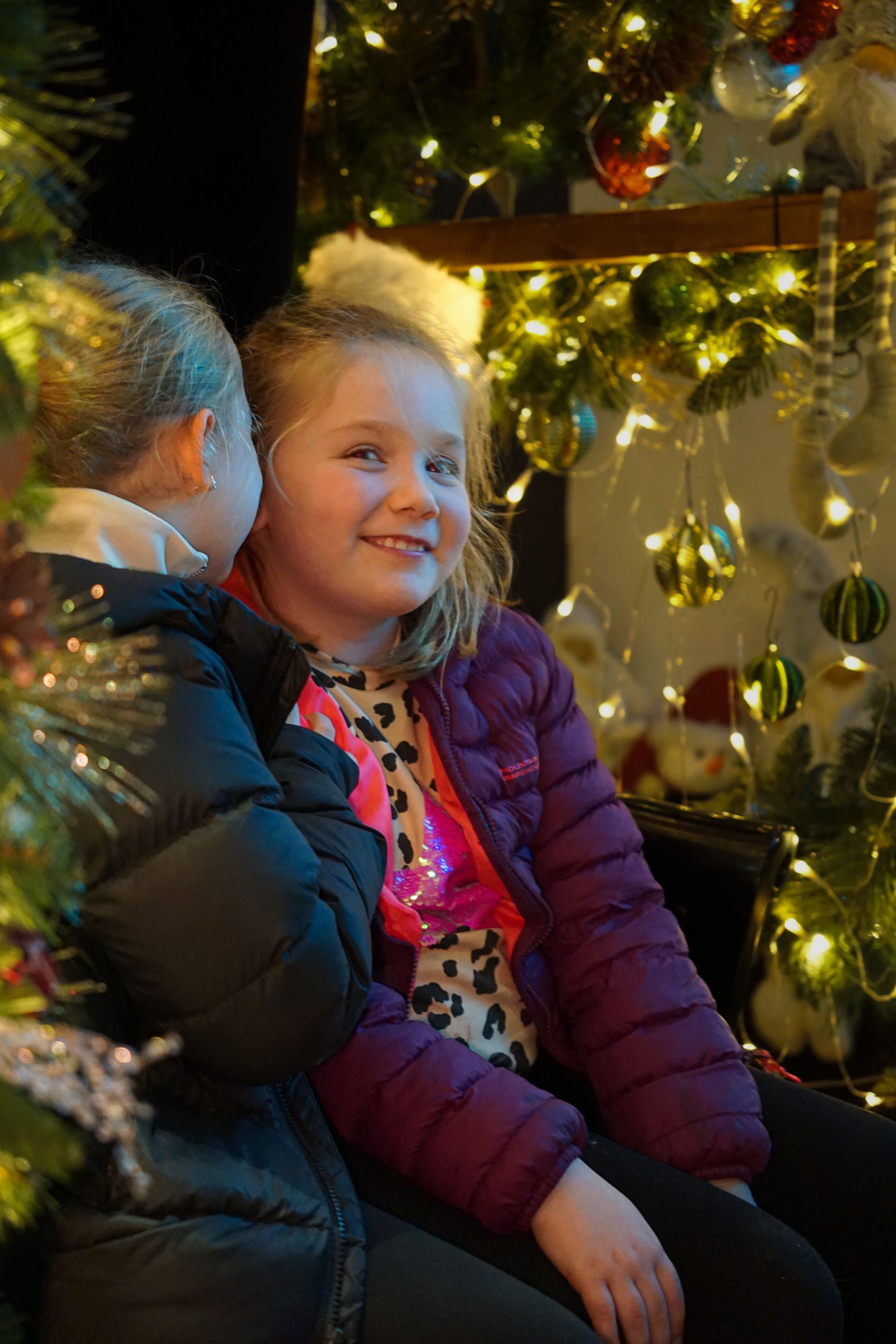 Two children in winter coats sitting beside a lit Christmas tree, smiling in warm holiday lights at Stump Cross Caverns