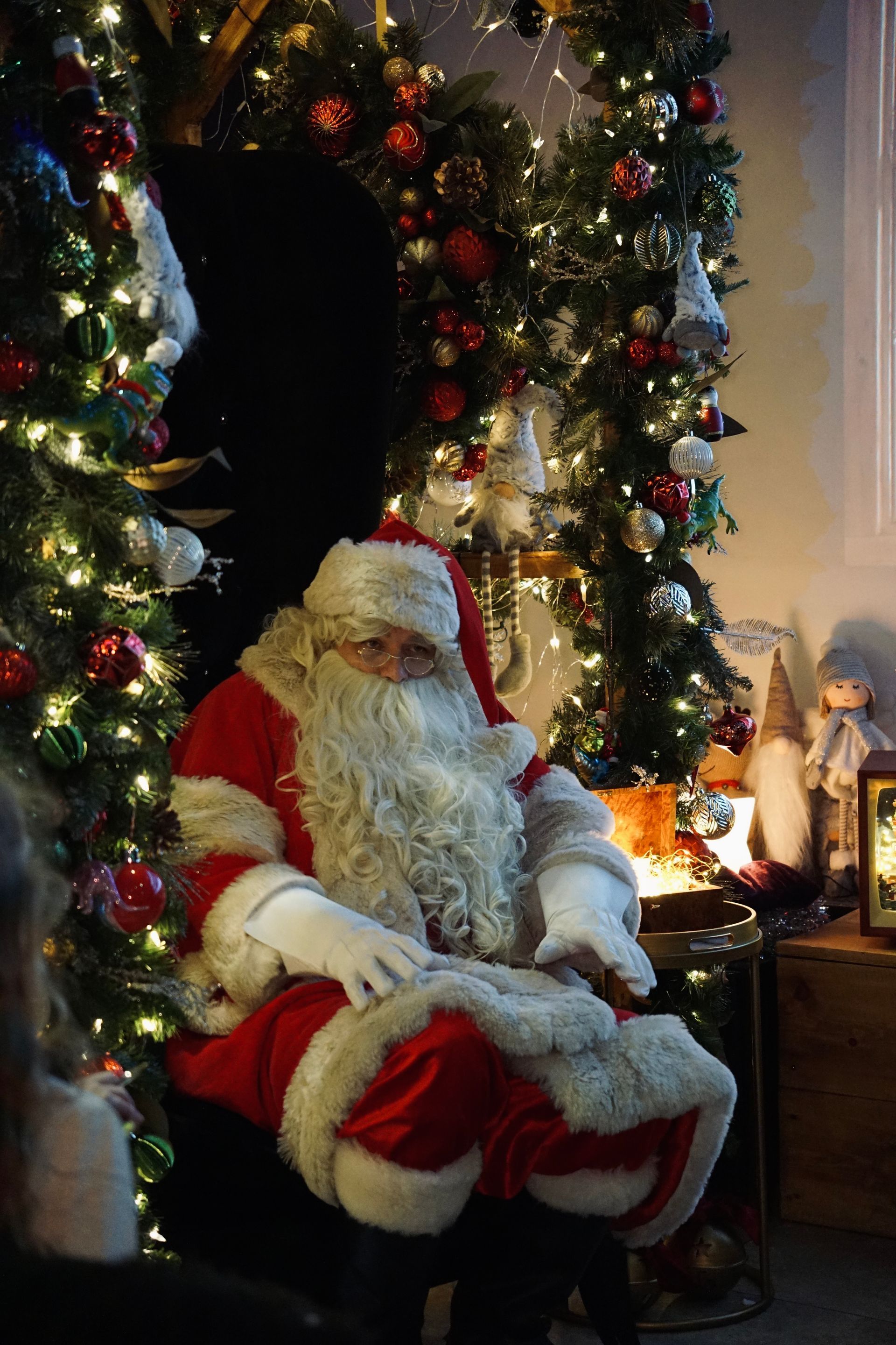 Santa Claus seated beside a decorated Christmas tree in a cozy, festive room