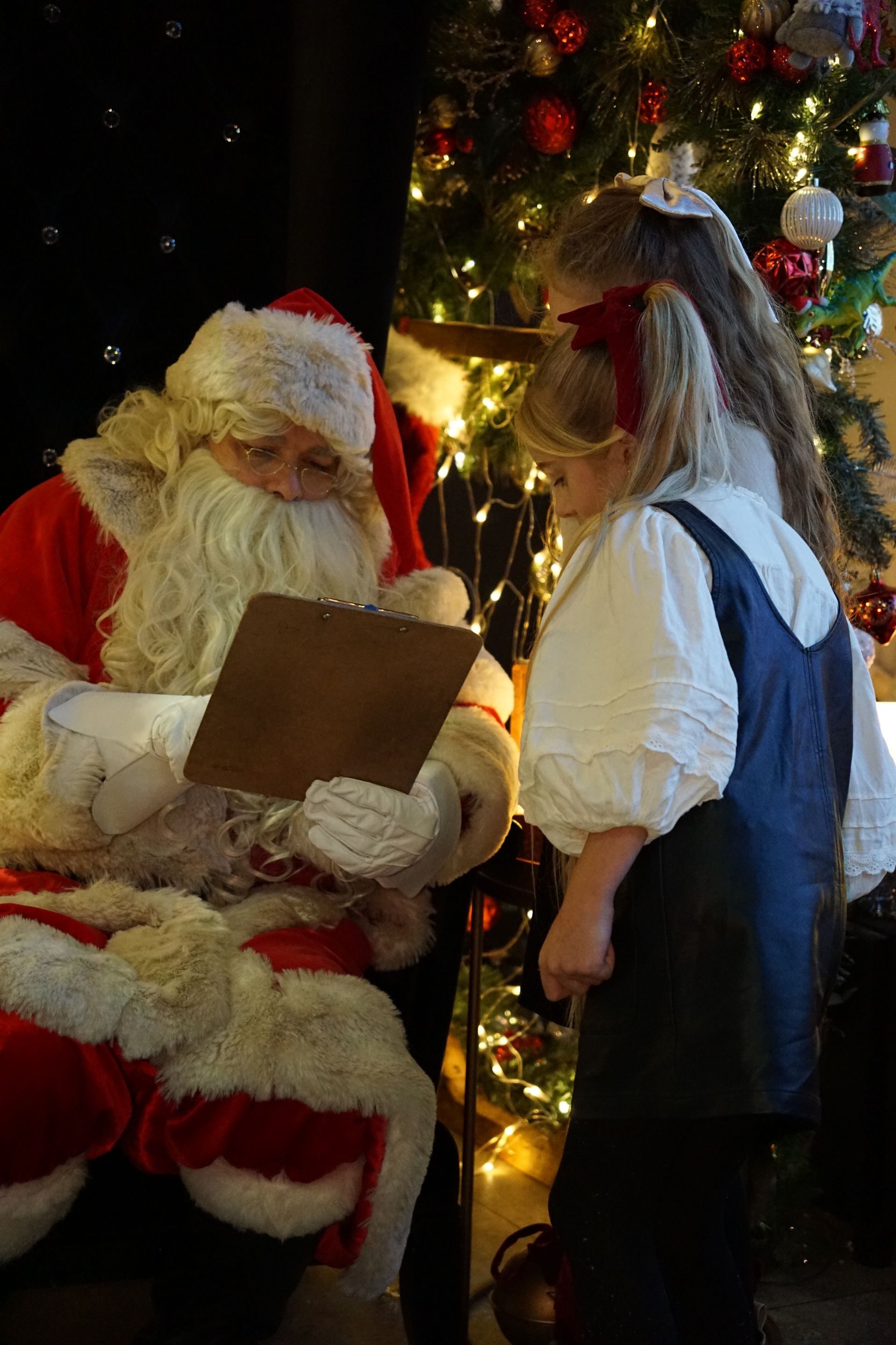 Santa seated beside a decorated Christmas tree, reading a clipboard to a child in costume at Stump Cross Caverns