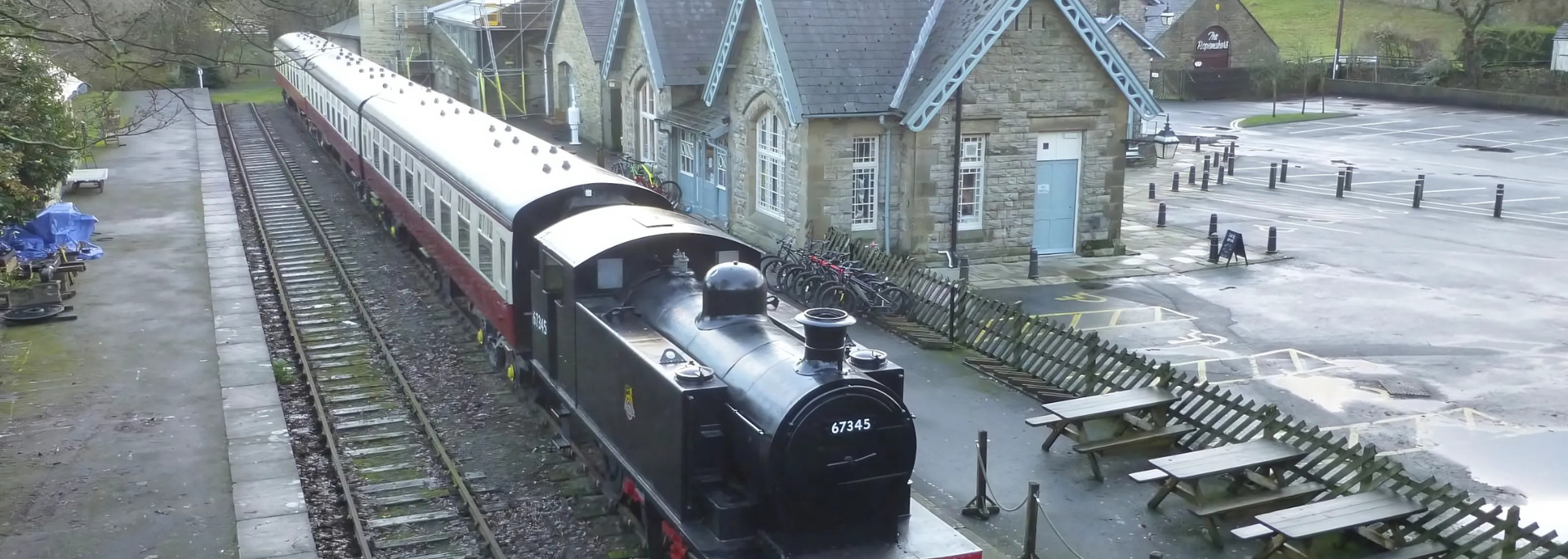 Steam train at a station with stone buildings and picnic tables.