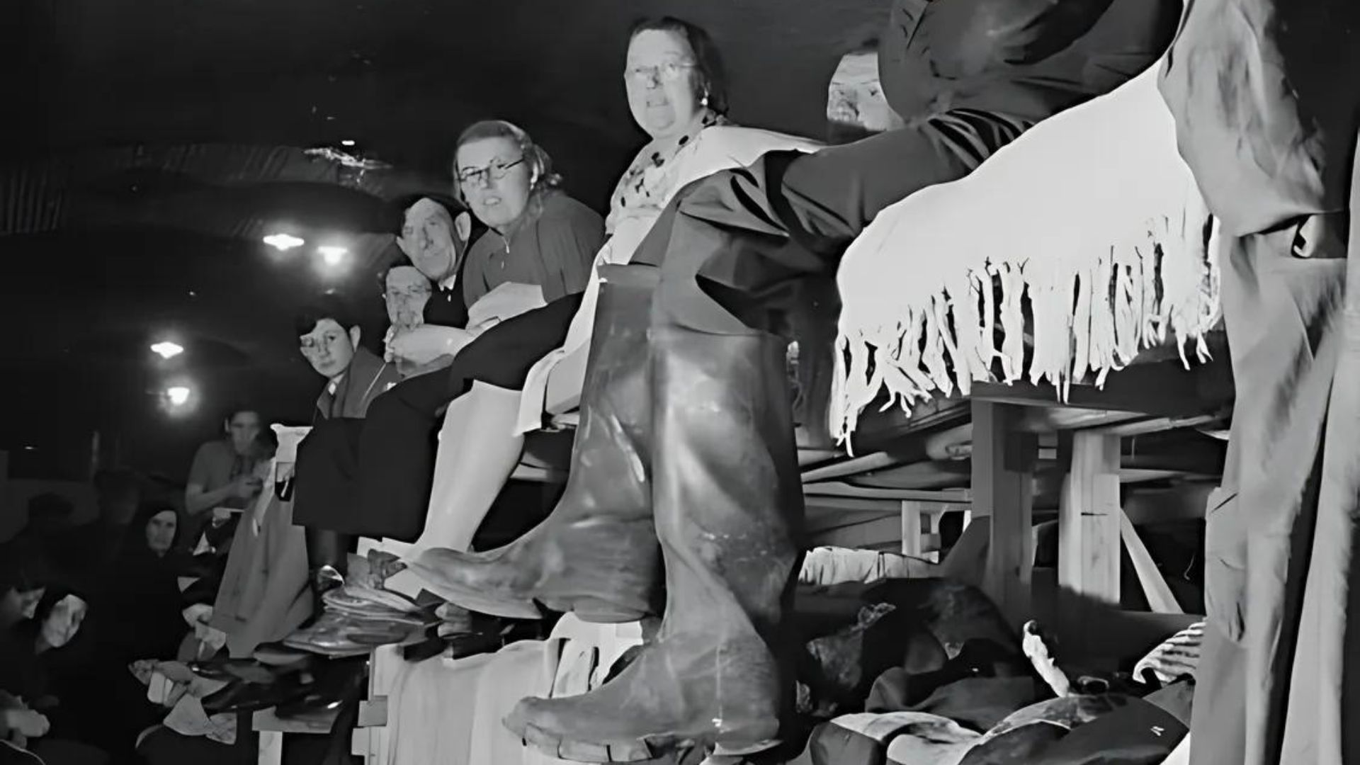 People seated, some looking at large boots on display. Indoor setting, likely a market.