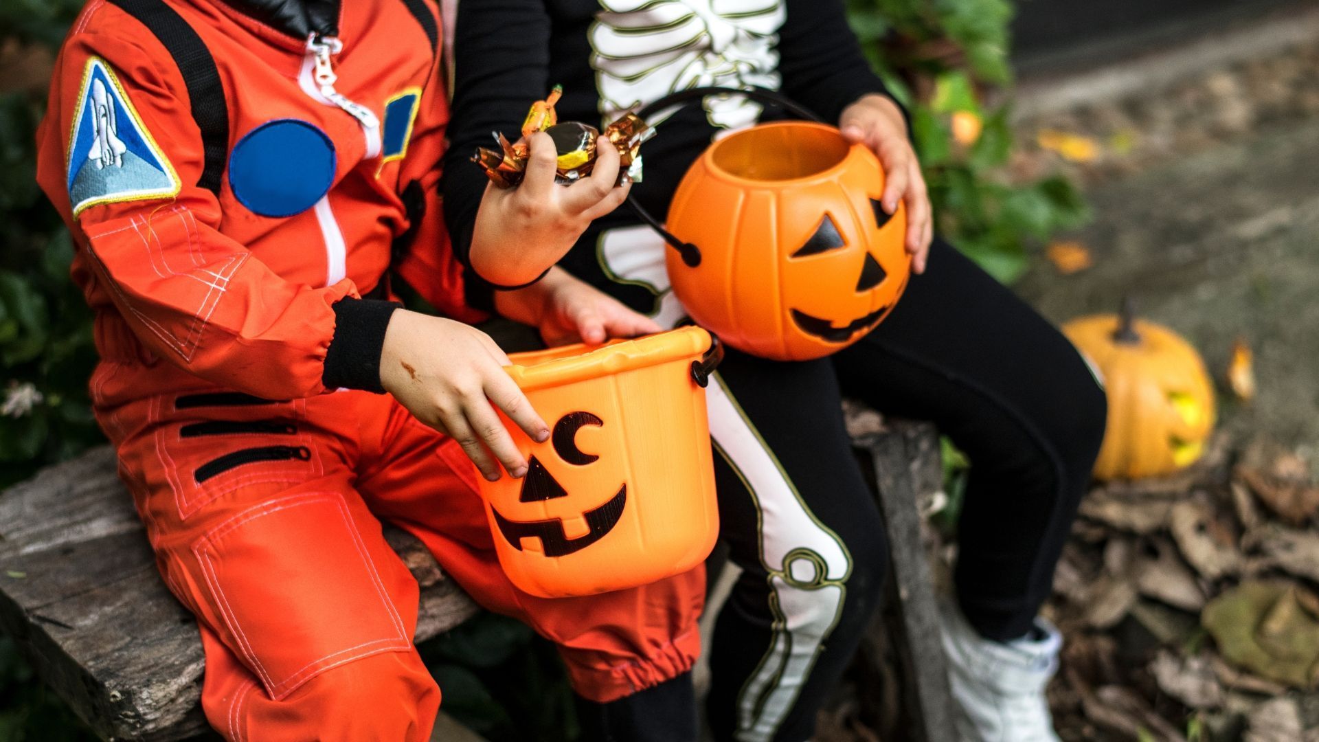 Two children in Halloween costumes sit with pumpkin-shaped candy buckets. One wears an astronaut suit, the other a skeleton costume.