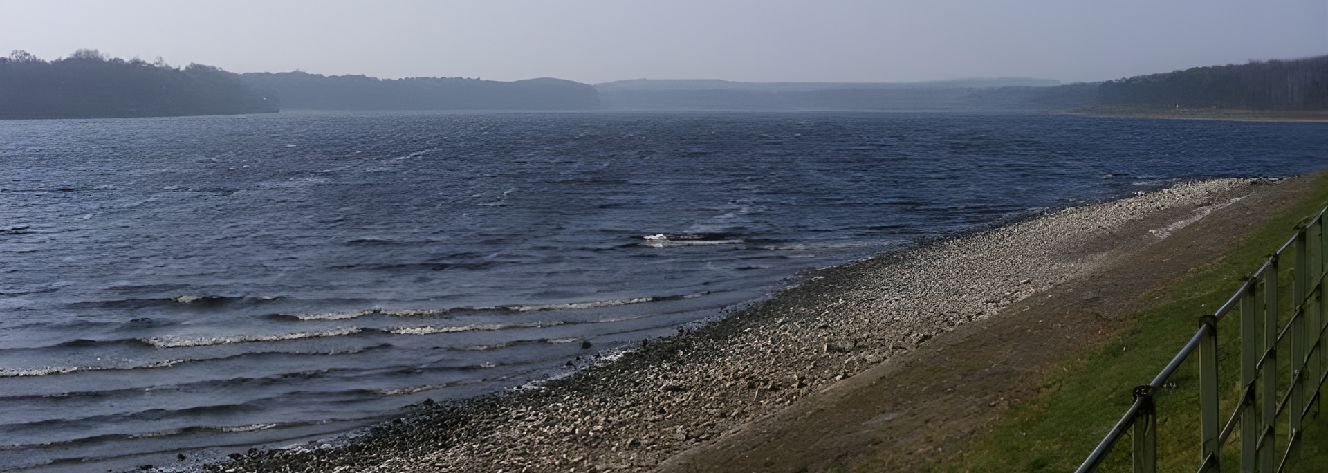 Watery lake with a rocky shore and treed distant banks under a pale sky.