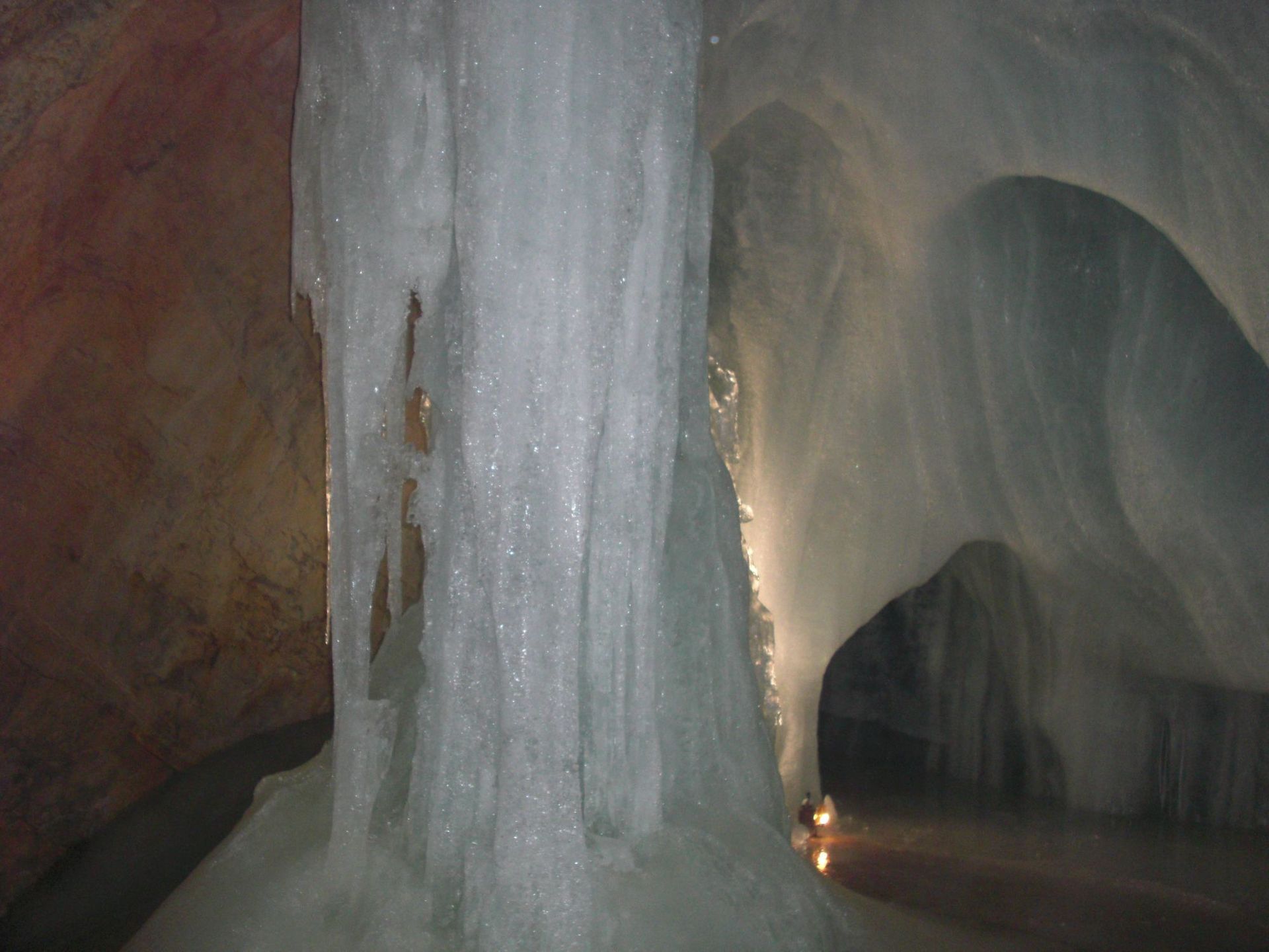 Inside an ice cave, a large, cylindrical ice formation stands center, with light reflecting off the ice walls.
