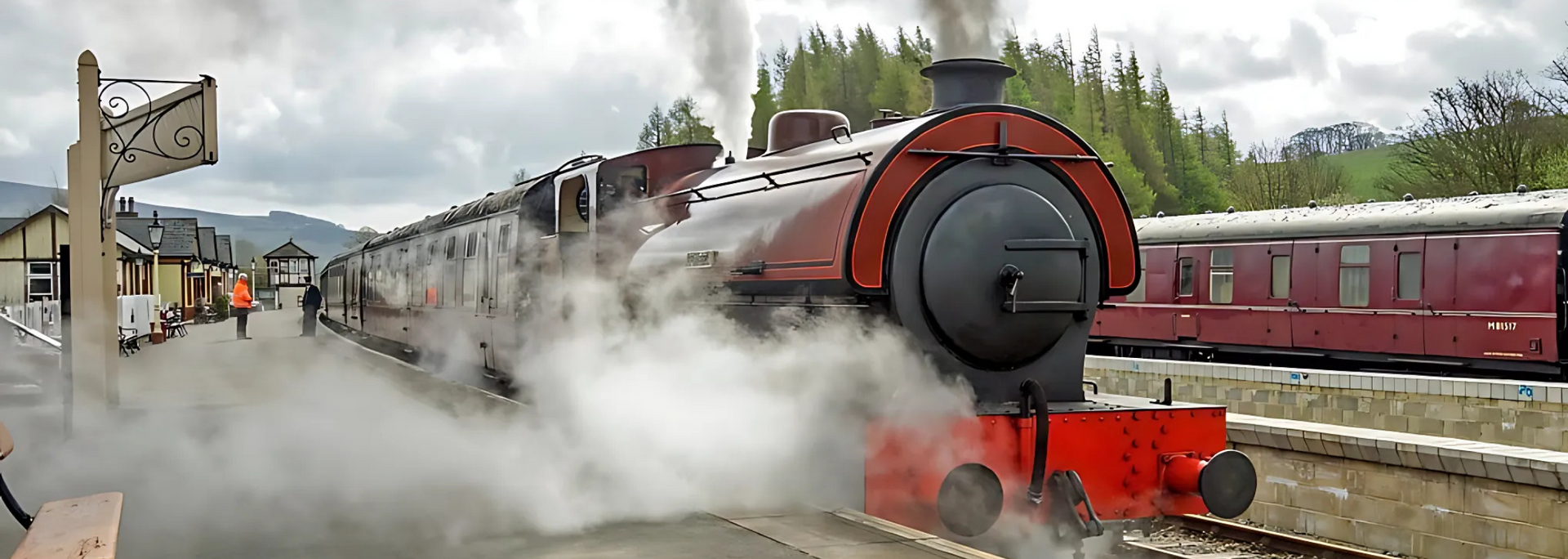 Steam train at a station. Maroon and black engine with billowing white steam. Other maroon cars nearby.