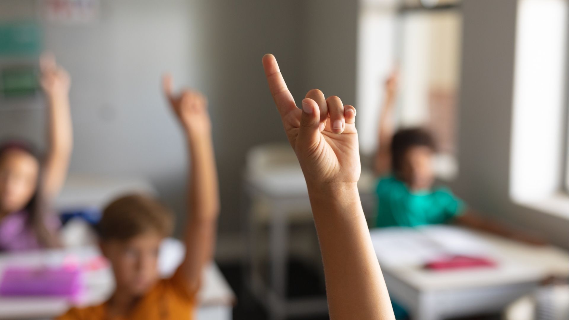 Hands raised in a classroom, one finger pointed up prominently.