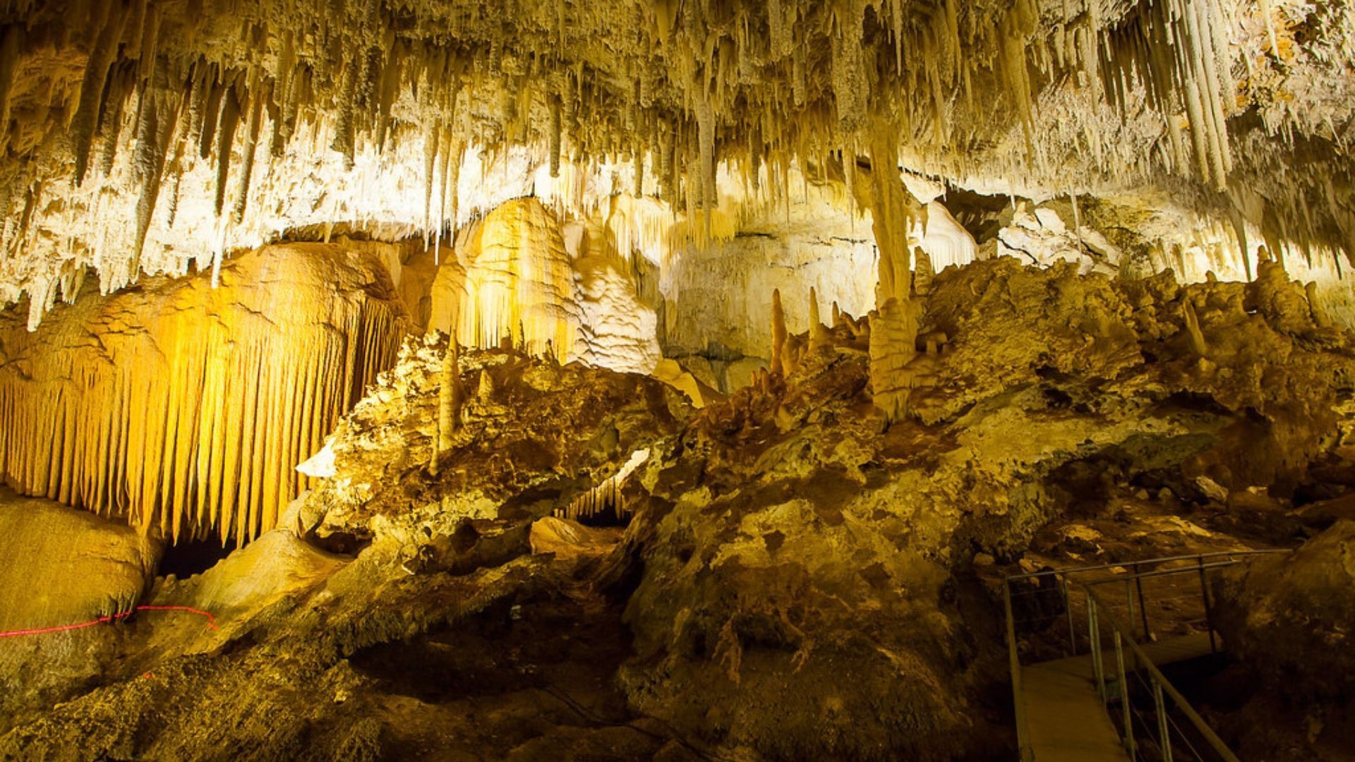 A cave interior with golden stalactites and stalagmites. Sunlight illuminates the formations, a staircase is visible.