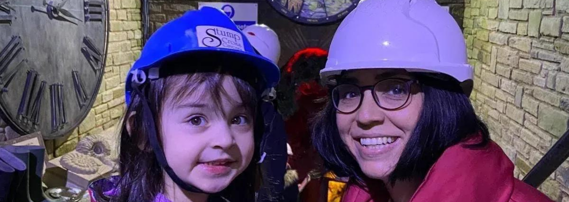 Two people smiling, wearing hard hats, in a room with a large clock and stone walls.