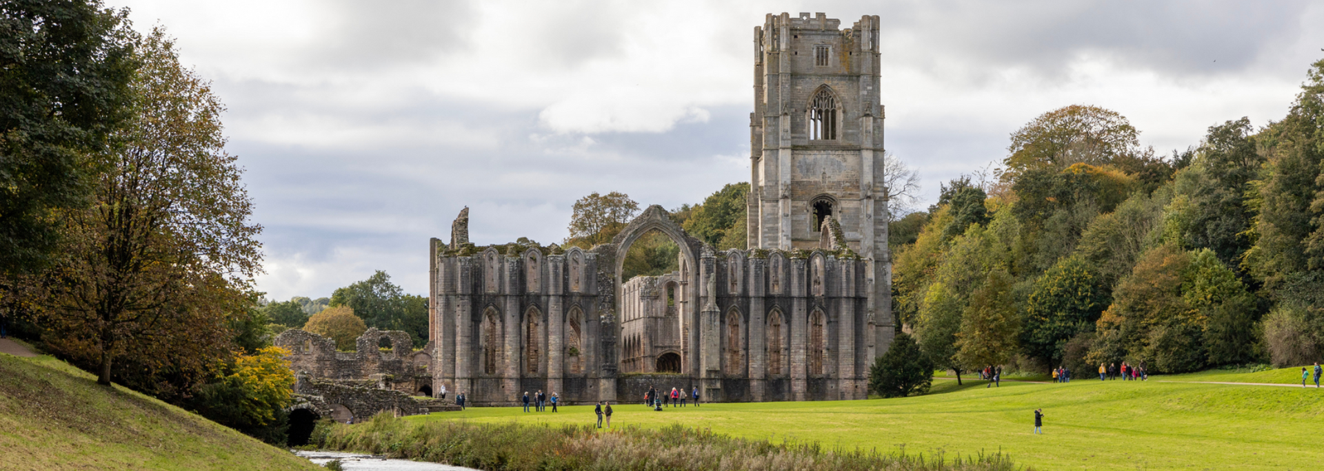Ruins of a stone abbey on a green lawn with trees and a cloudy sky.