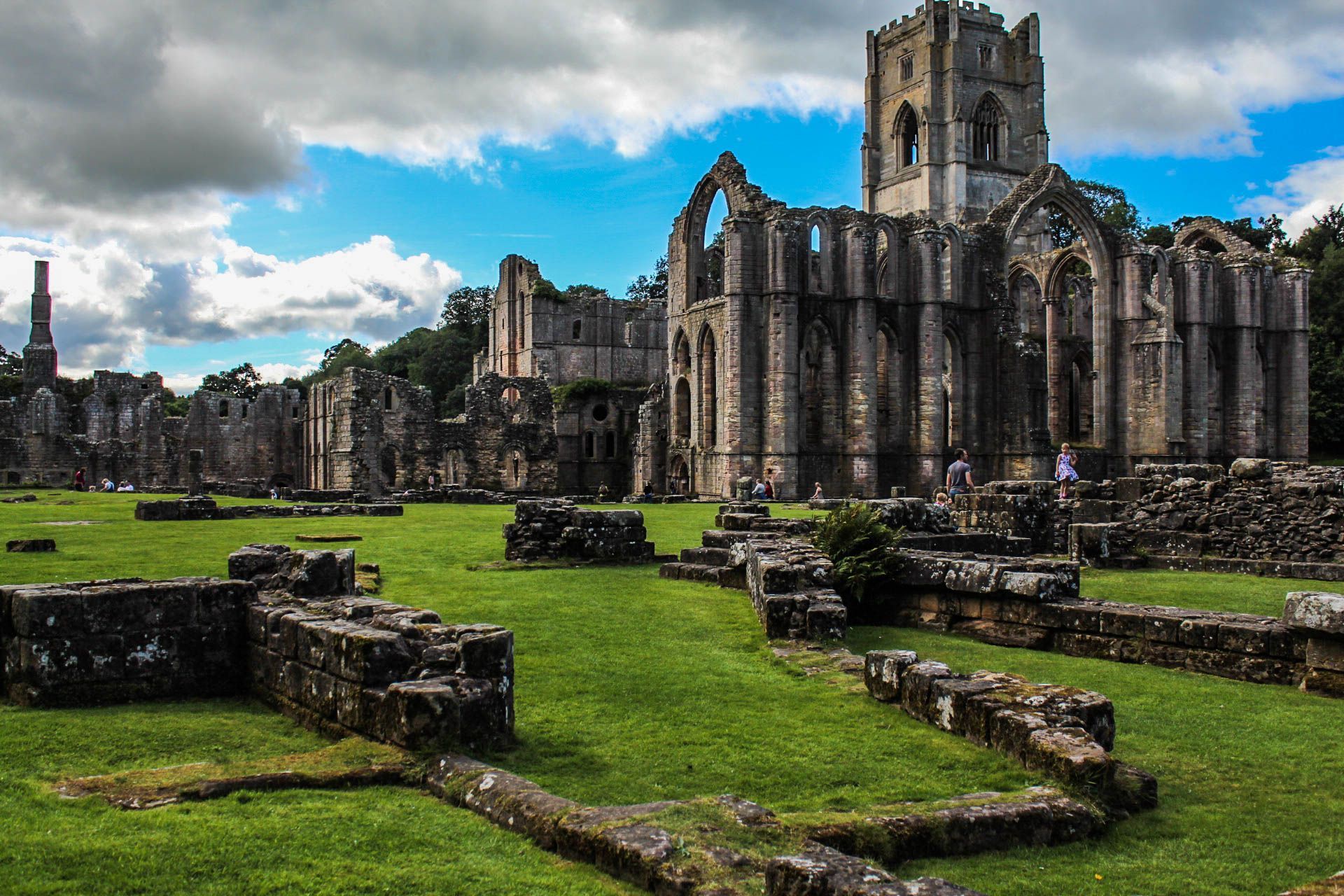 Ruins of Fountains Abbey in England, with a central tower and surrounding stone structures in a grassy field.