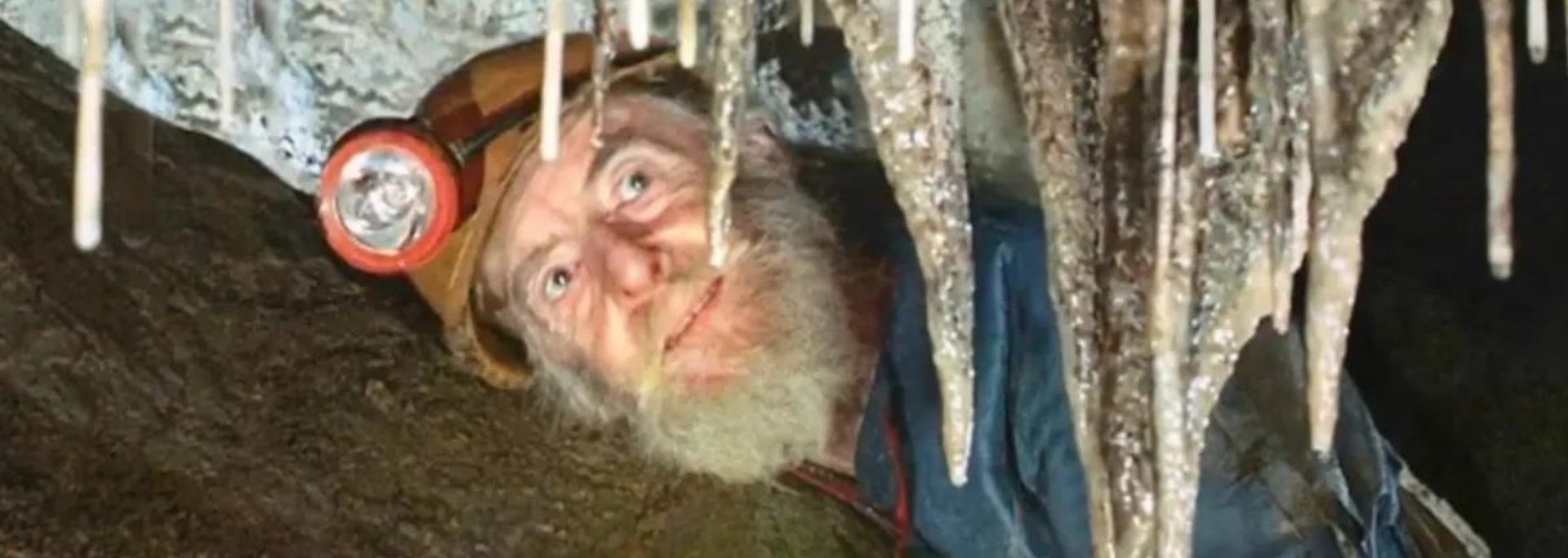 A man wearing a headlamp smiles while looking up at stalactites in a cave.