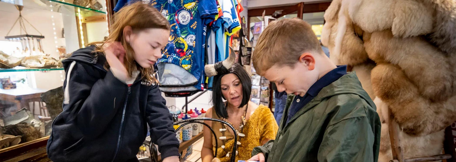 Two children and a woman examining items in a shop. The children are focused on something in their hands.