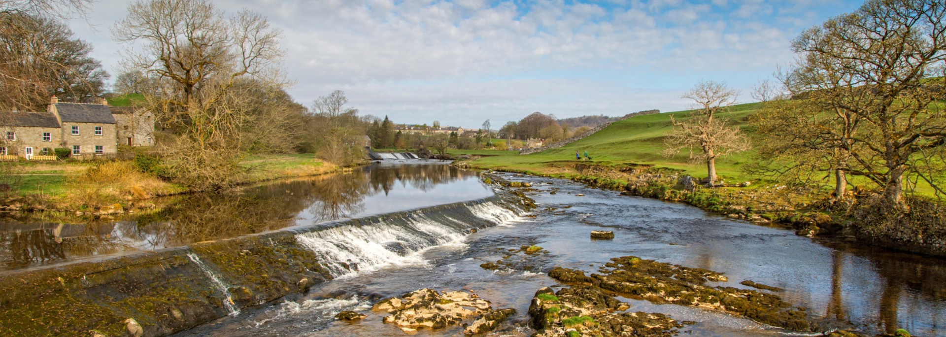 River flowing through a rural landscape with stone buildings and grassy banks.