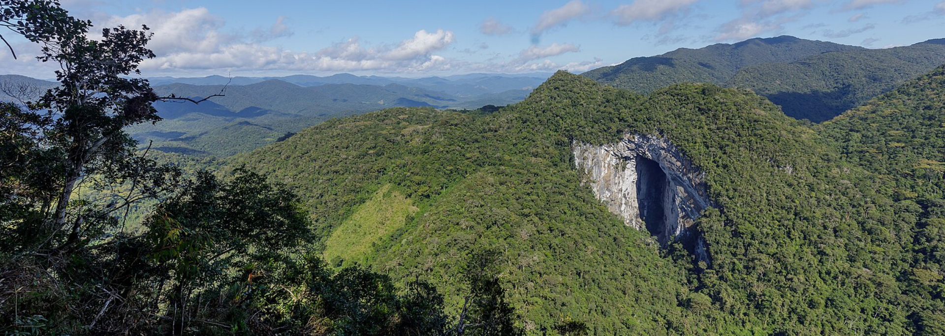 Panoramic view of lush green mountains under a bright, cloudy sky, with a large, cave-like opening in the hillside.