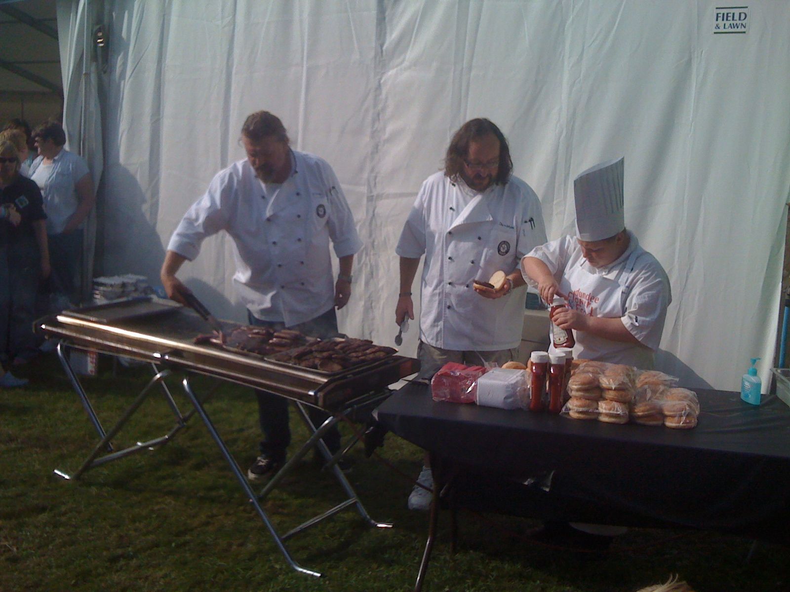 Chefs grilling burgers at an outdoor event, one preparing buns with condiments.
