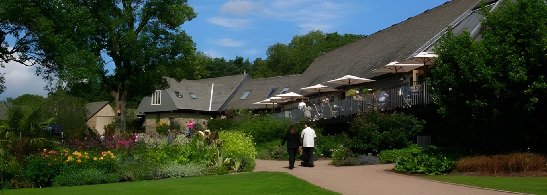 Building with outdoor seating, surrounded by greenery and two people on a pathway.