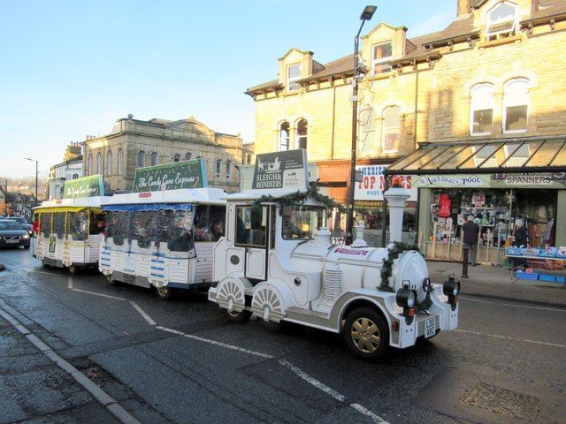 White tourist train on a road in front of brick buildings, people visible inside carriages.