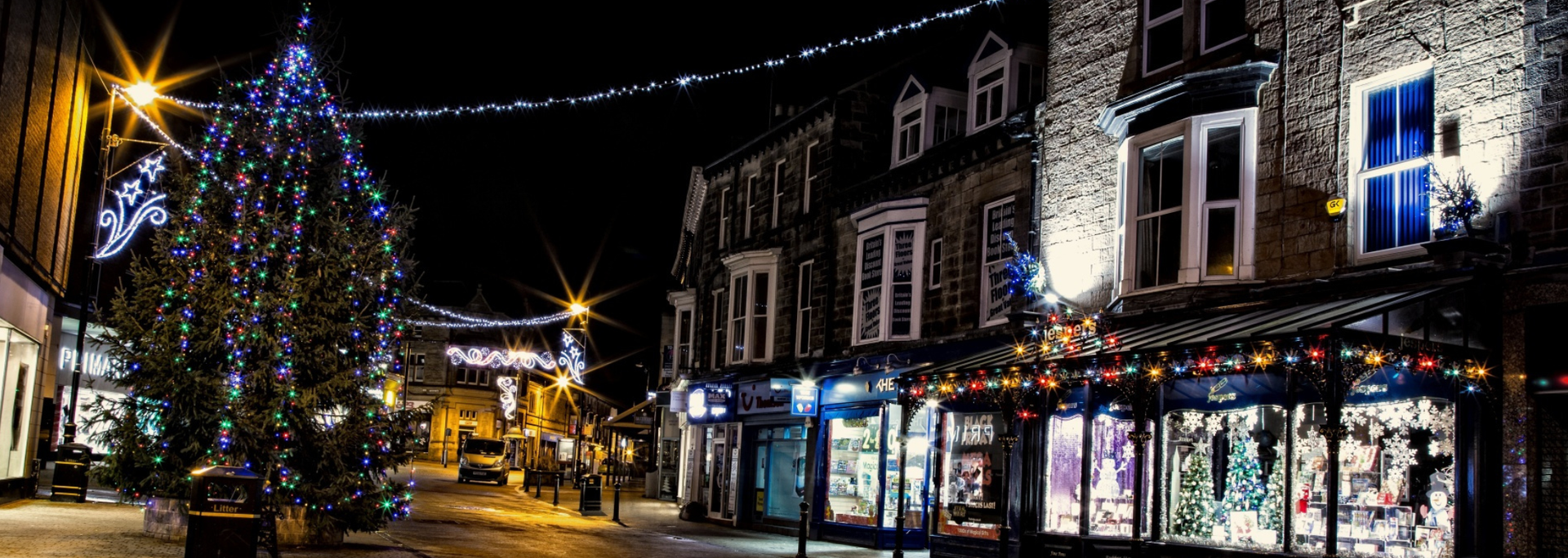 Nighttime street scene decorated for Christmas with shops, lights, and a tree.