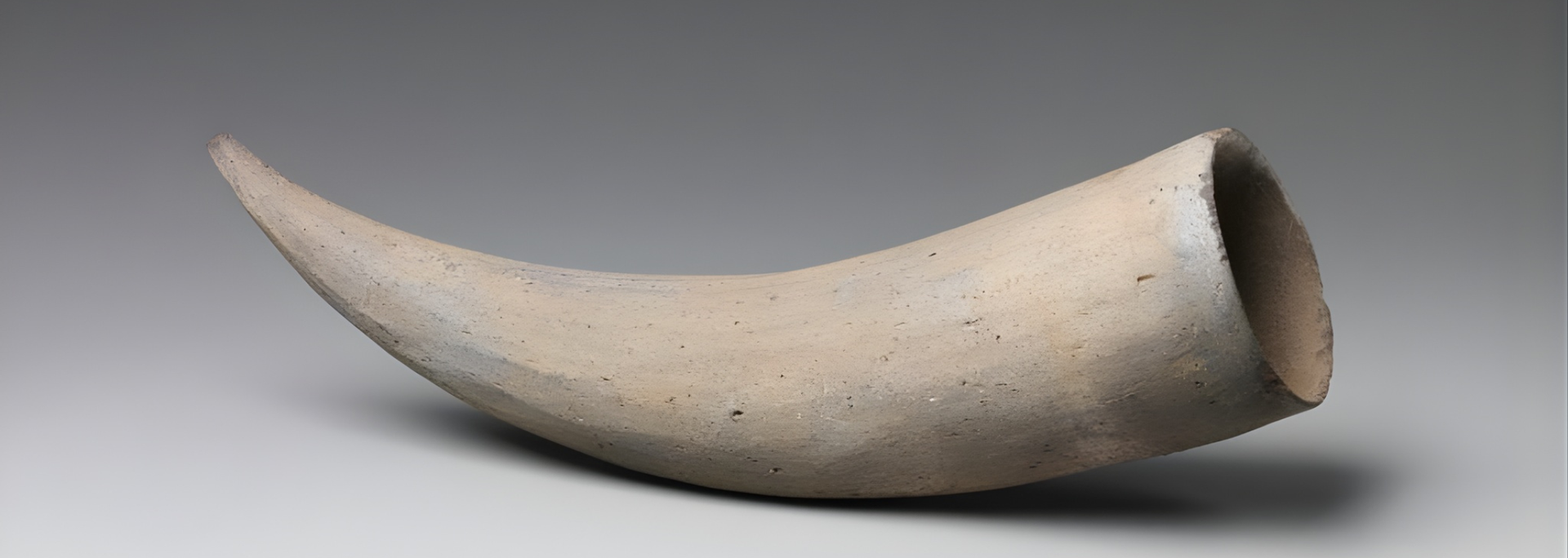 A curved, horn-shaped pottery vessel with a wide mouth. It is light tan against a grey background.