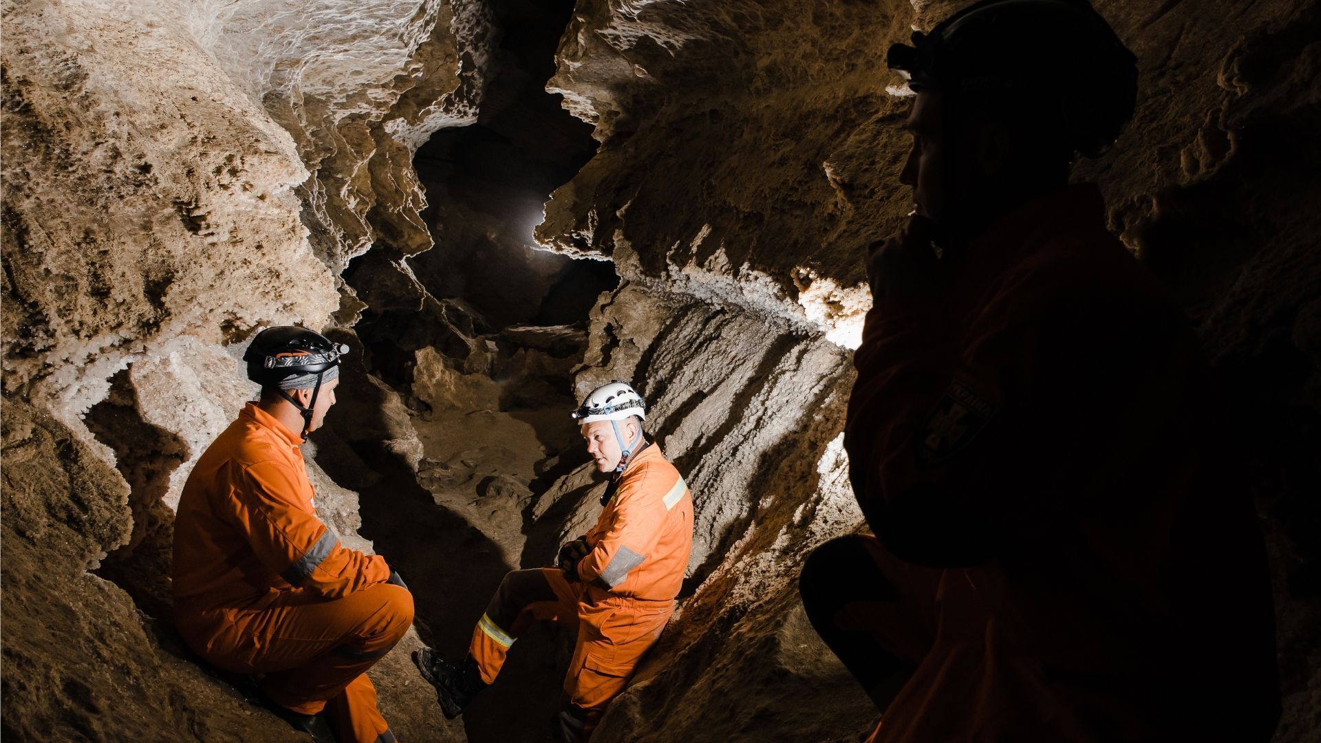 Three people in orange suits and helmets explore a cave, illuminated by headlamps.