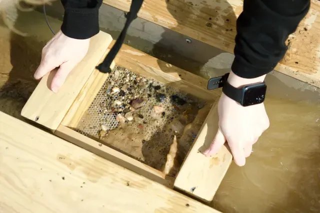 Person using a gold pan in water, sifting through gravel.