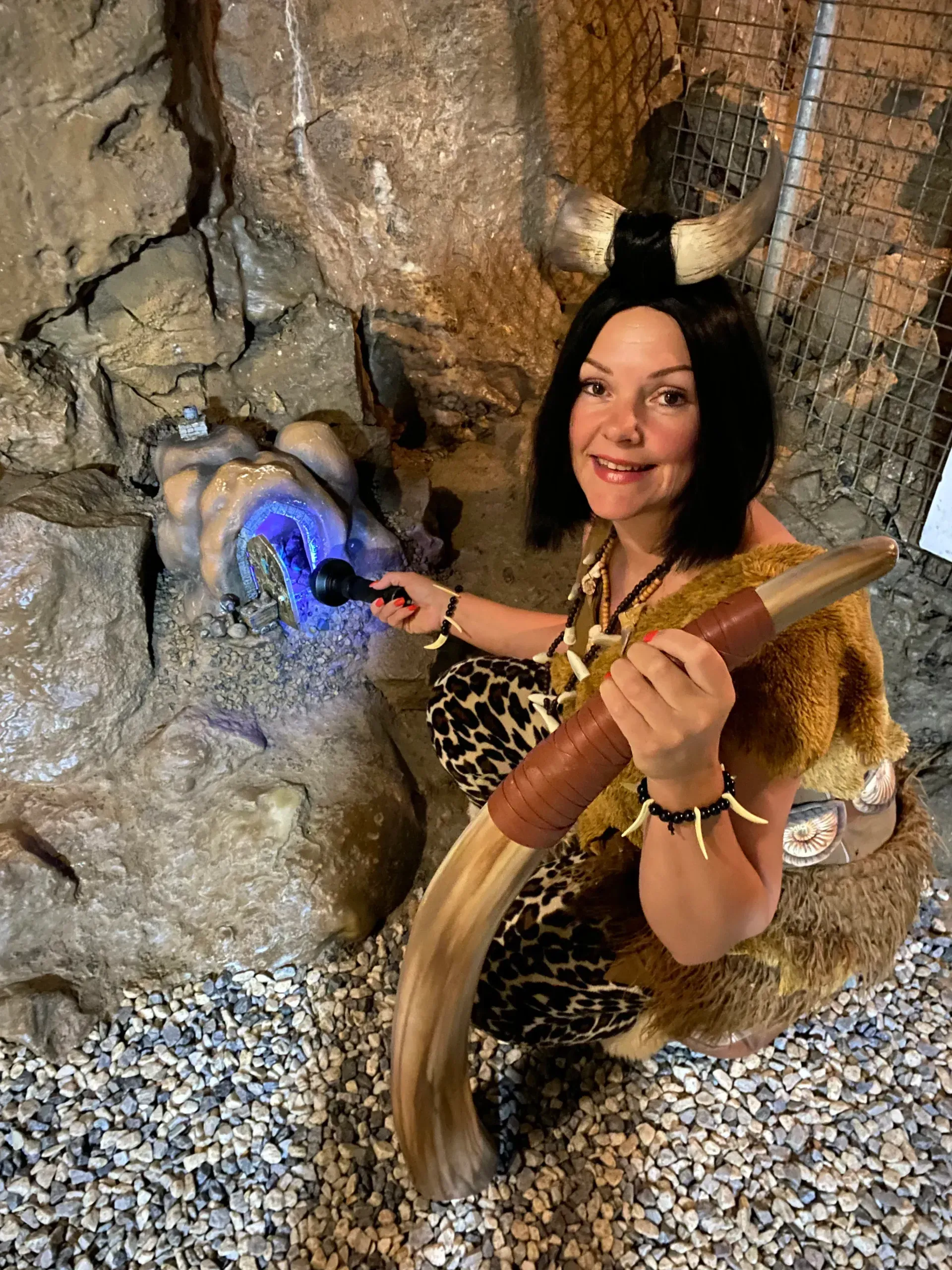 Woman in cave attire holding a staff, interacting with a glowing blue rock formation.