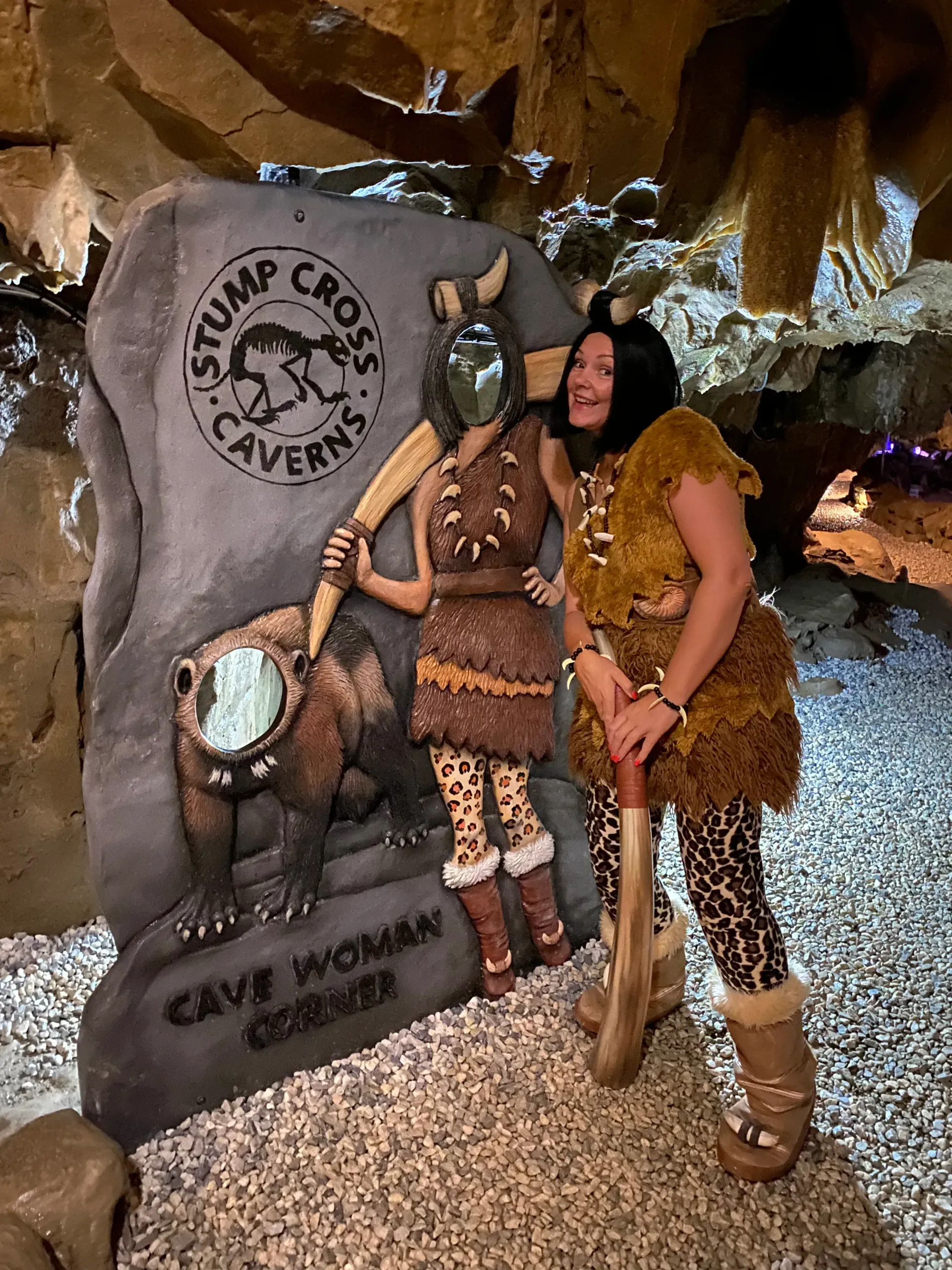 Woman in cave-woman costume smiles beside a sign for Stump Cross Caverns with a cutout for photos.
