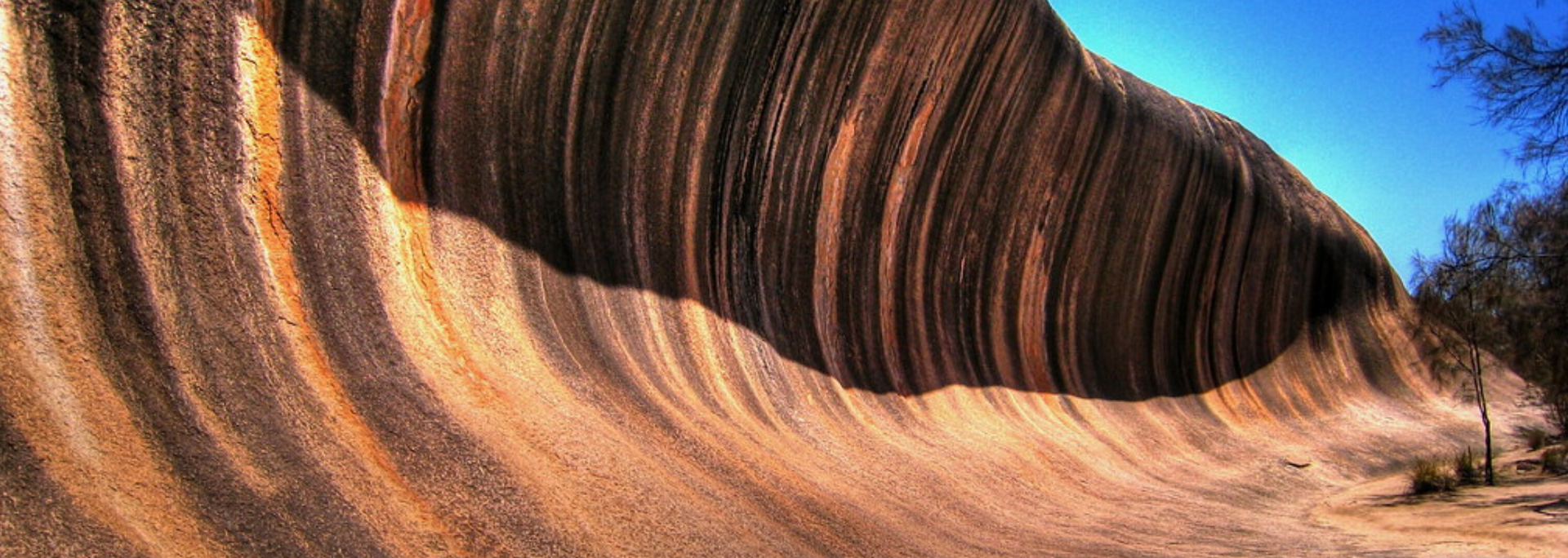 Wave Rock, a natural rock formation in Australia, with brown and tan striped surface under a blue sky.