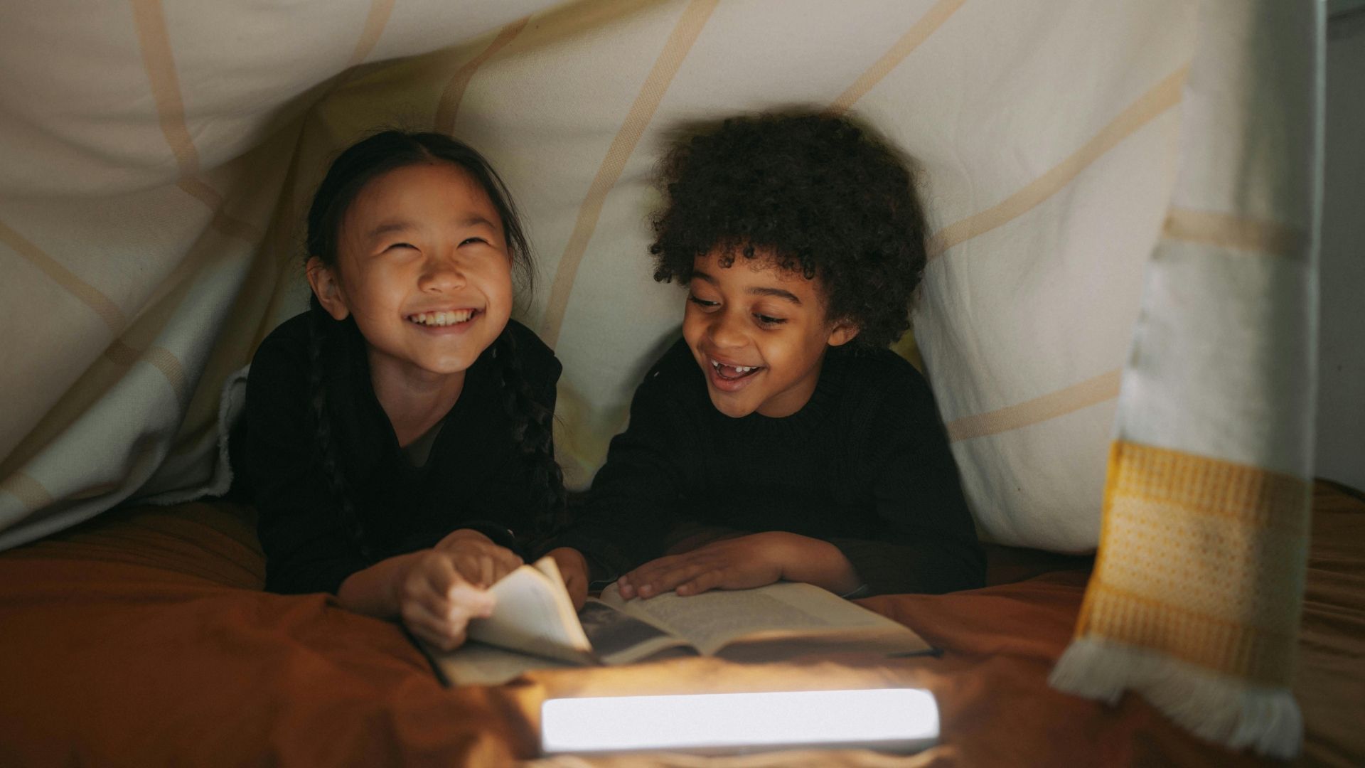Two children laugh while reading a book inside a blanket fort, lit by a lamp.