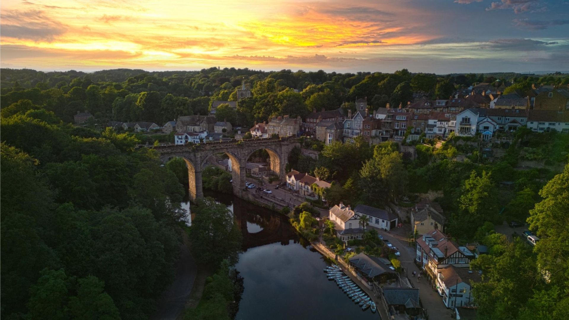 Sunset over Knaresborough, England; stone viaduct arches over a river. Trees and buildings frame the scene.