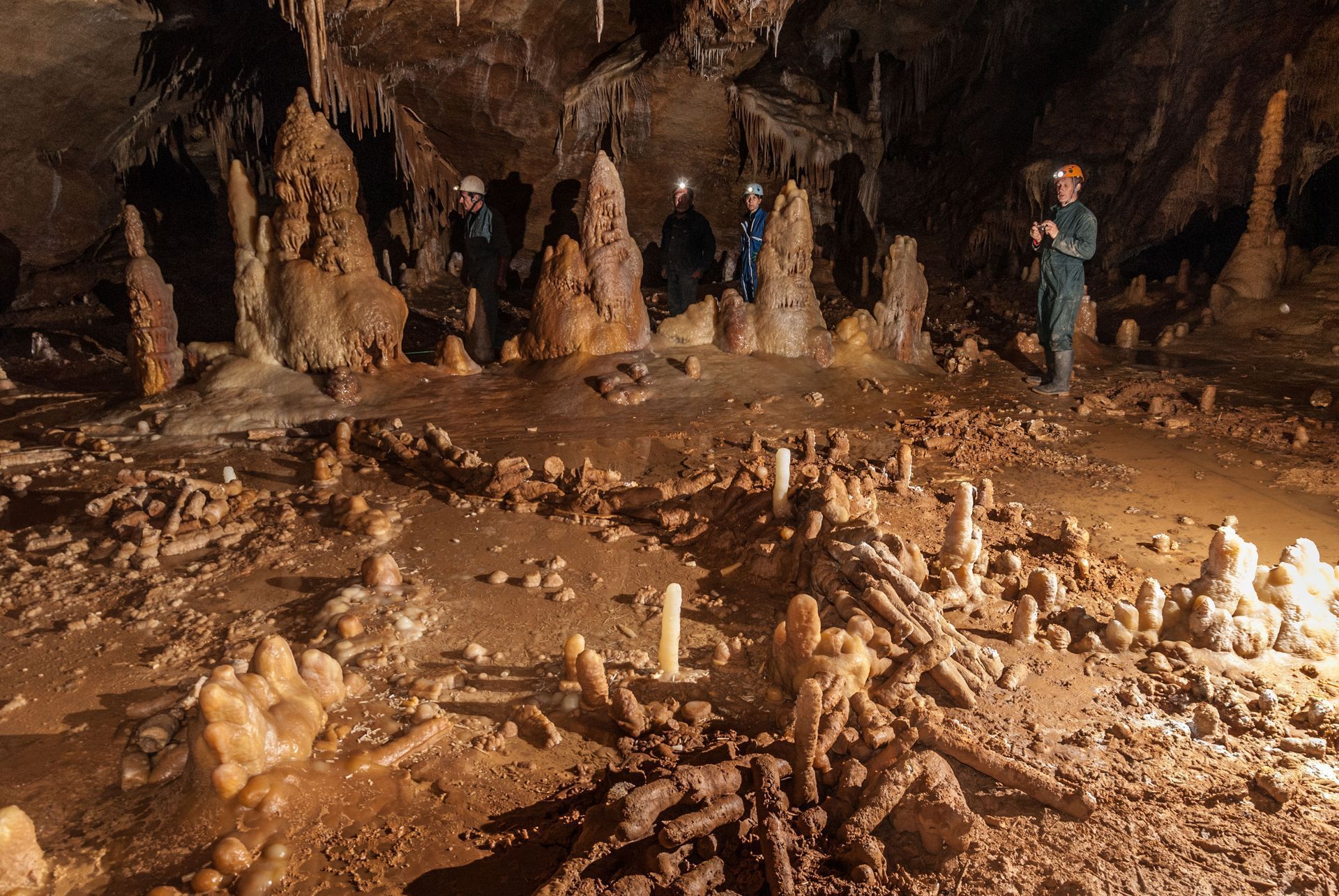 People exploring a cave with stalagmites and stalactites. They wear headlamps and protective gear.