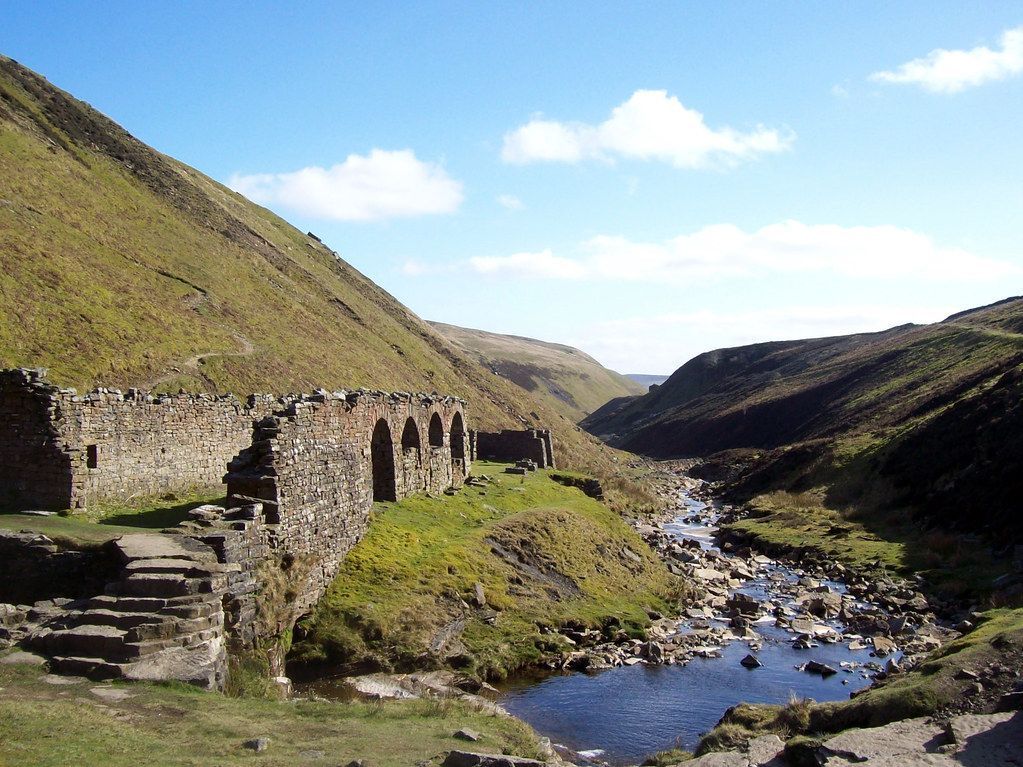 Stone ruins next to a river running through a valley under a blue sky.