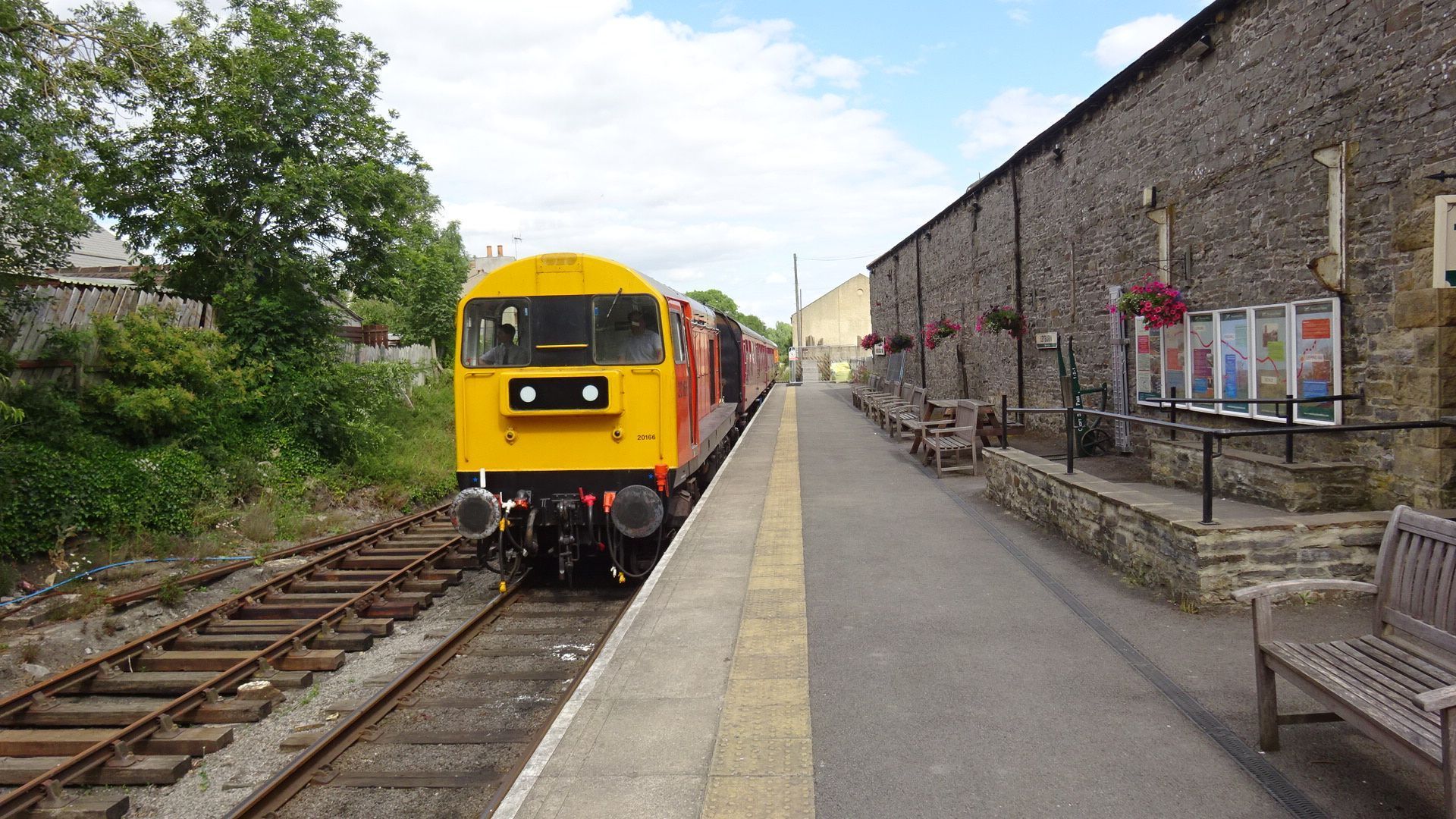 Yellow and red train at a train station platform with a stone building on the right.