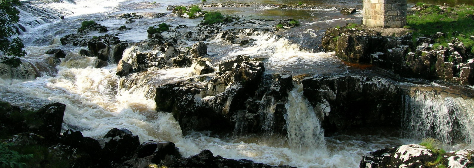 Rushing water cascading over dark, rocky terrain, creating a small waterfall. Green vegetation lines the banks.