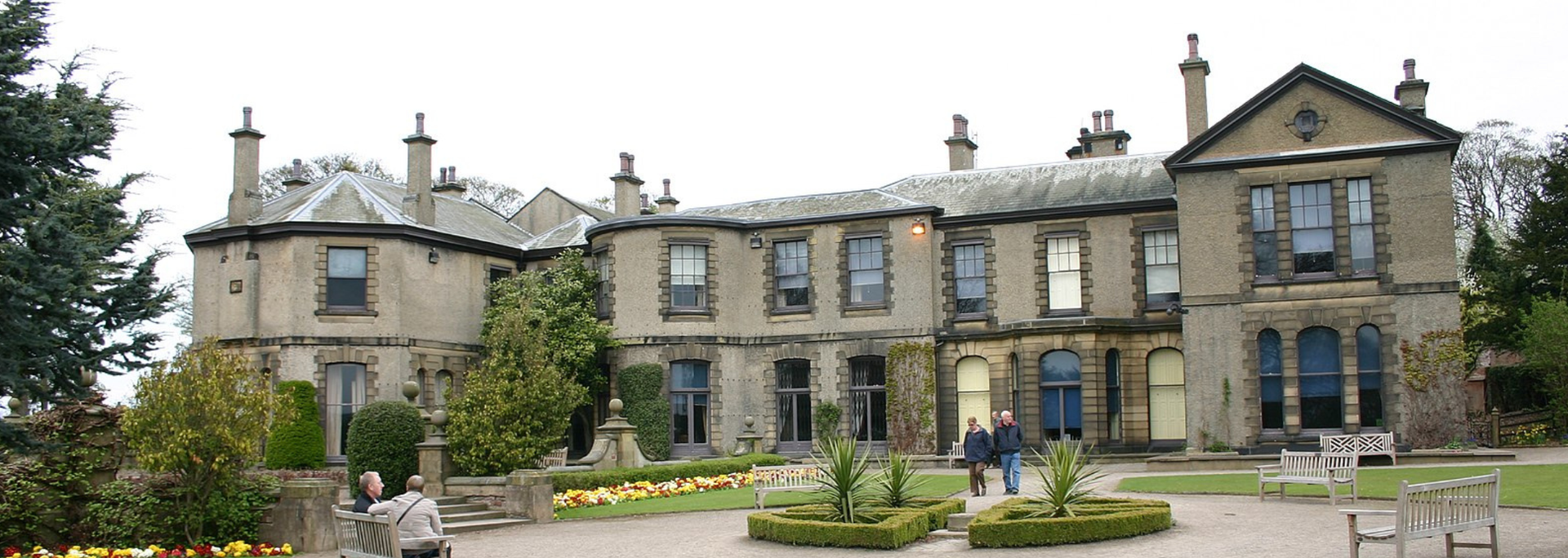 Large stone building with multiple windows, surrounded by a garden and trees.